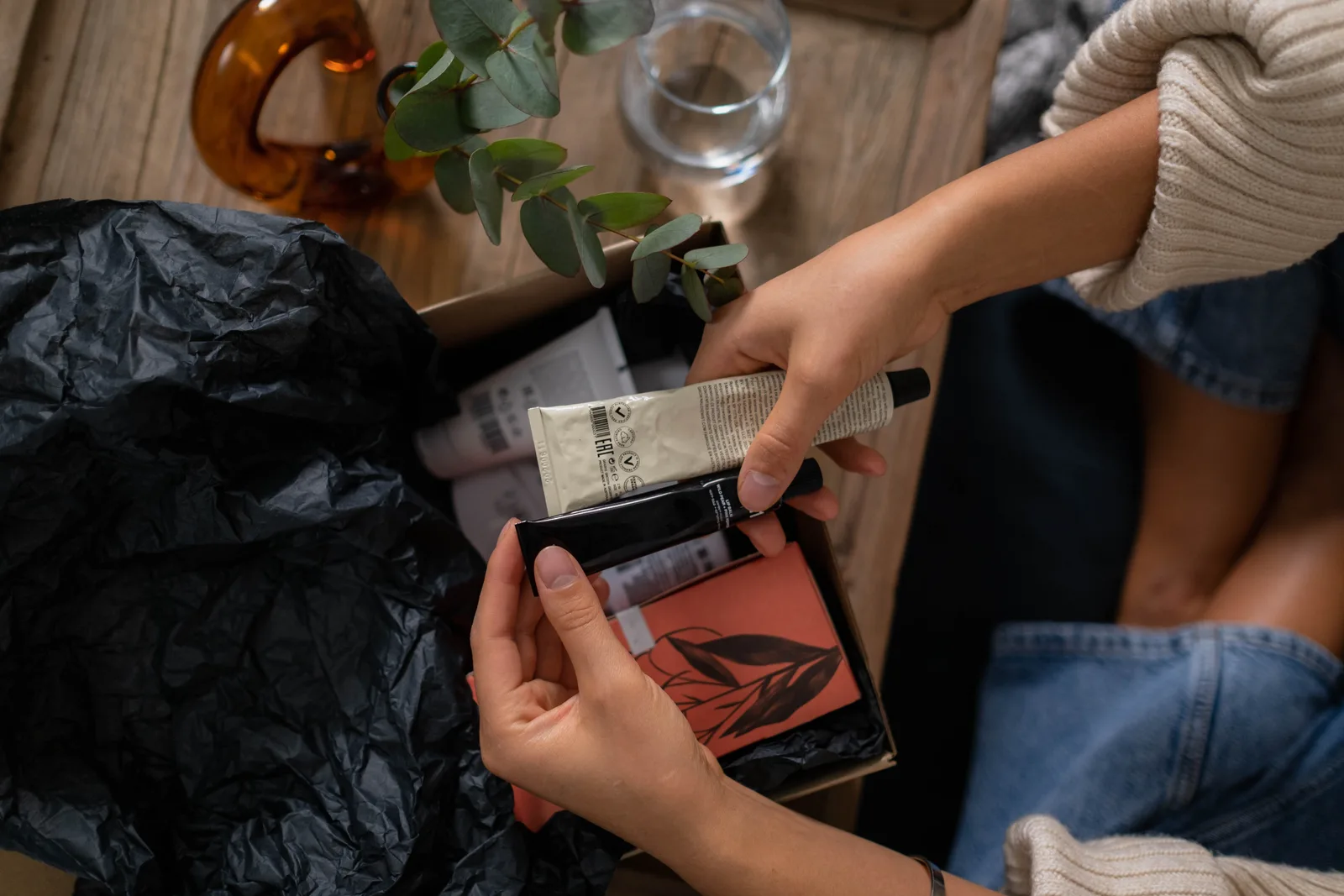 Hands unboxing skincare products with eucalyptus on wooden surface.